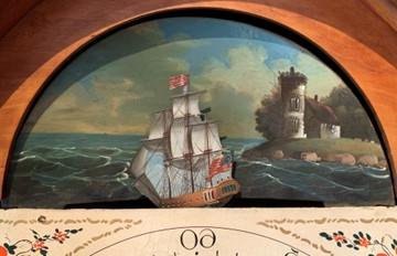 Color photo showing a painted seascape in a lunette above the dial face of a tall case clock. The painting shows a stone building on a grassy outcrop with rocks at the shoreline. In front of the scenery is a ship (three-masted frigate).