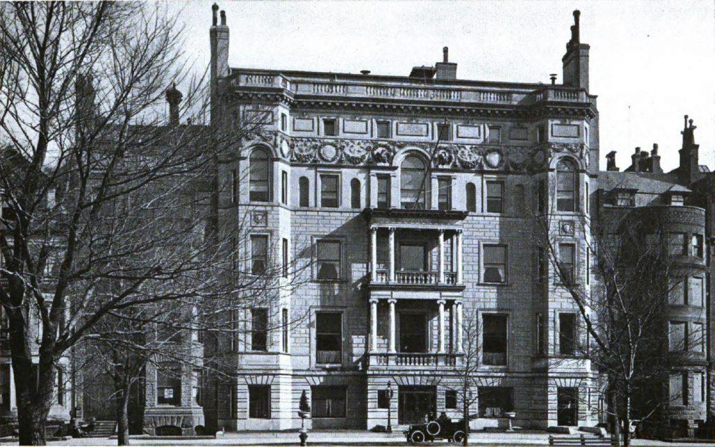 Black and white photograph of a 5-story building. The building has ornate decoration. On floors two and three, there is a balcony flanked by two columns in the middle of the facade. 